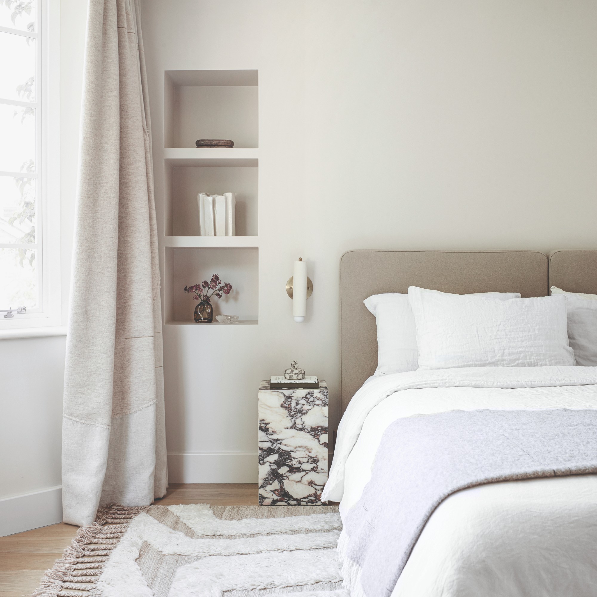 An off white-painted bedroom with a marble column bedside table, a tufted rug and a neutral upholstered headboard