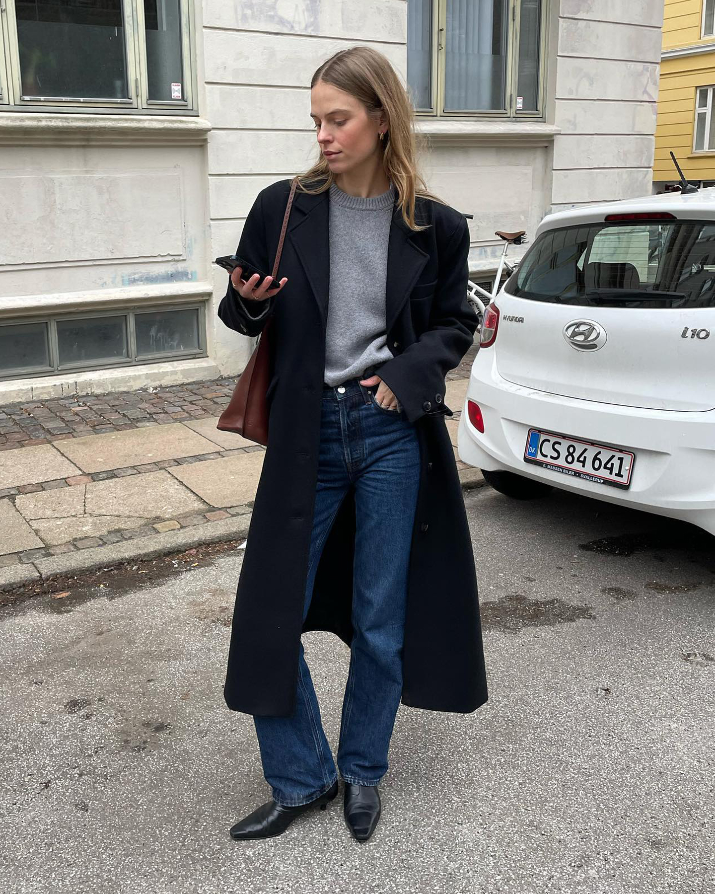 Scandinavian creative Clara Dyrhauge poses on the streets looking at her phone wearing a gray crewneck sweater, a long black coat, a brown shoulder bag, dark-wash straight-leg jeans, and black kitten-heel boots.