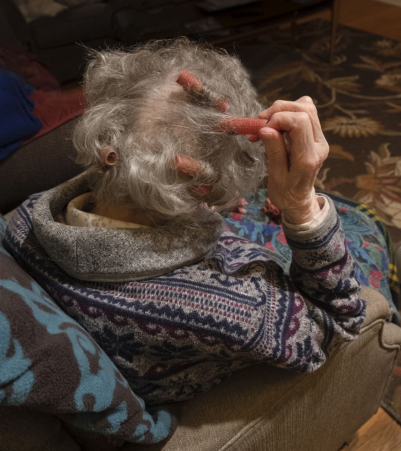 An elderly woman adjusts hair rollers in her curly gray hair while seated on a couch, wrapped in a patterned sweater