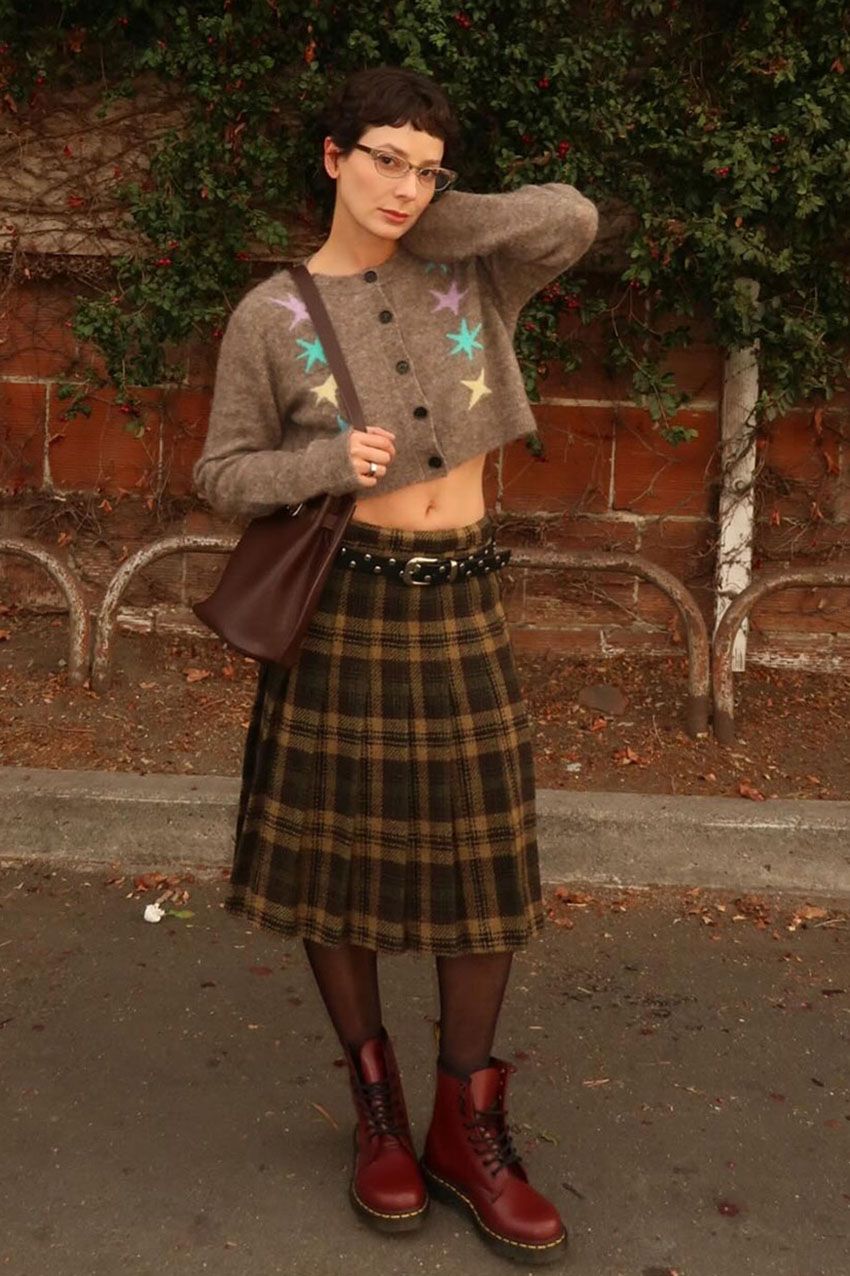A woman wearing a brown cardigan, plaid pleated skirt, and red boots