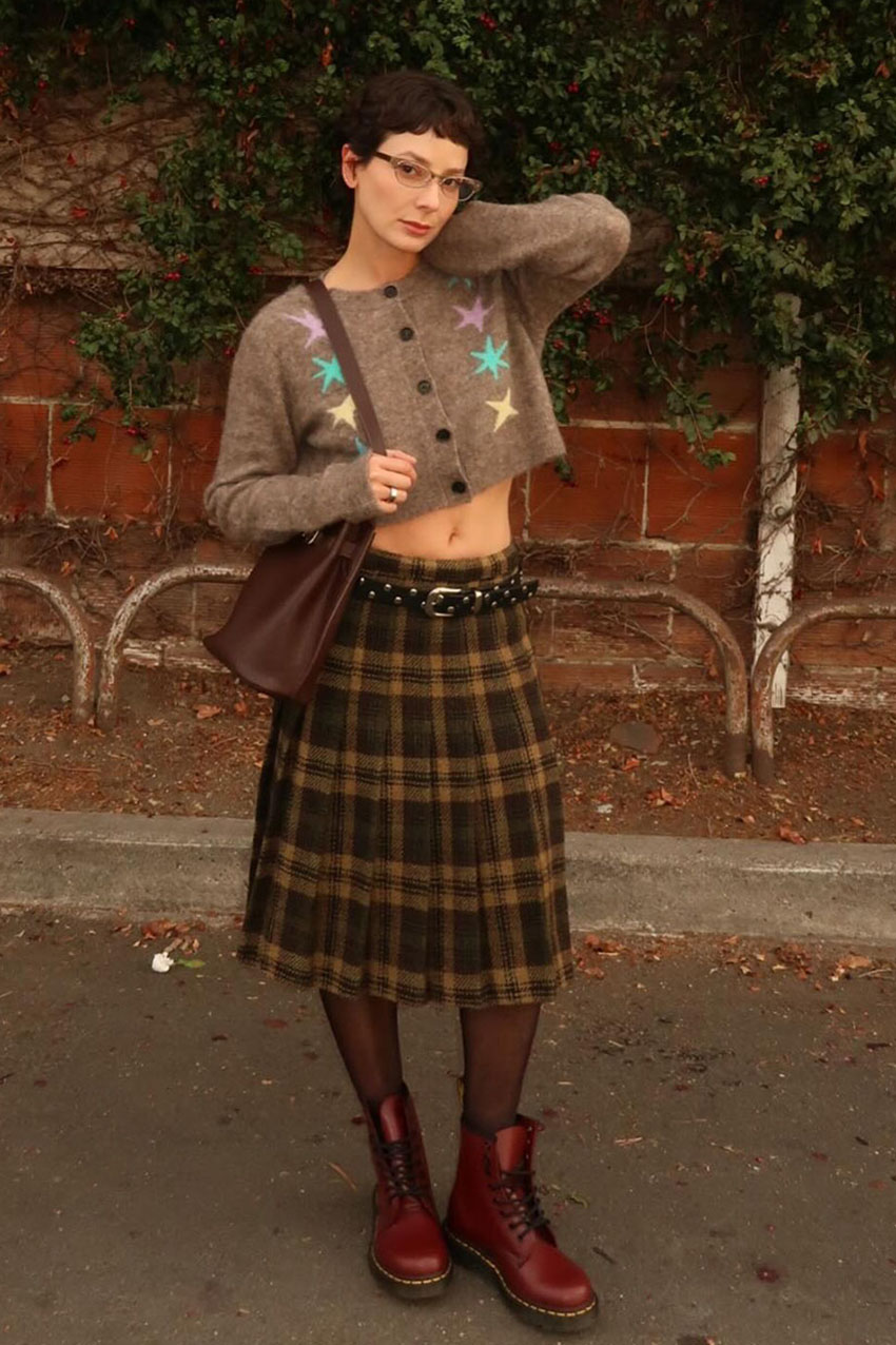 A woman wearing a brown cardigan, plaid pleated skirt, and red boots