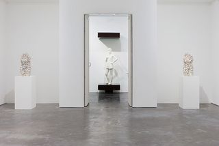 Two small statues are set on both sides of a door through which we see a white statue of a woman.