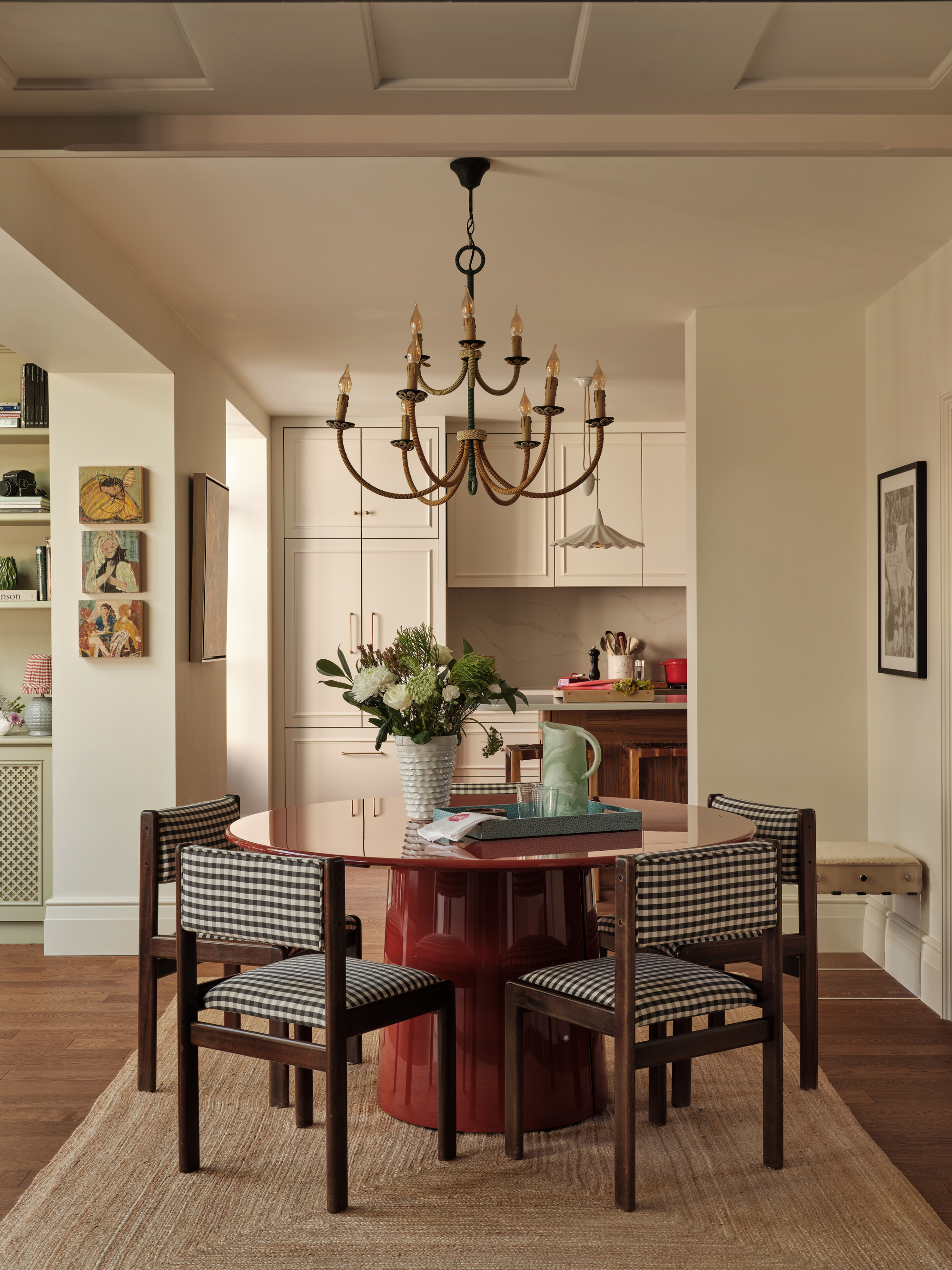 Image of a white dining room open to a white kitchen. There is a round, high-gloss, red dining table with a jute rug underneath and five black and white gingham chairs.