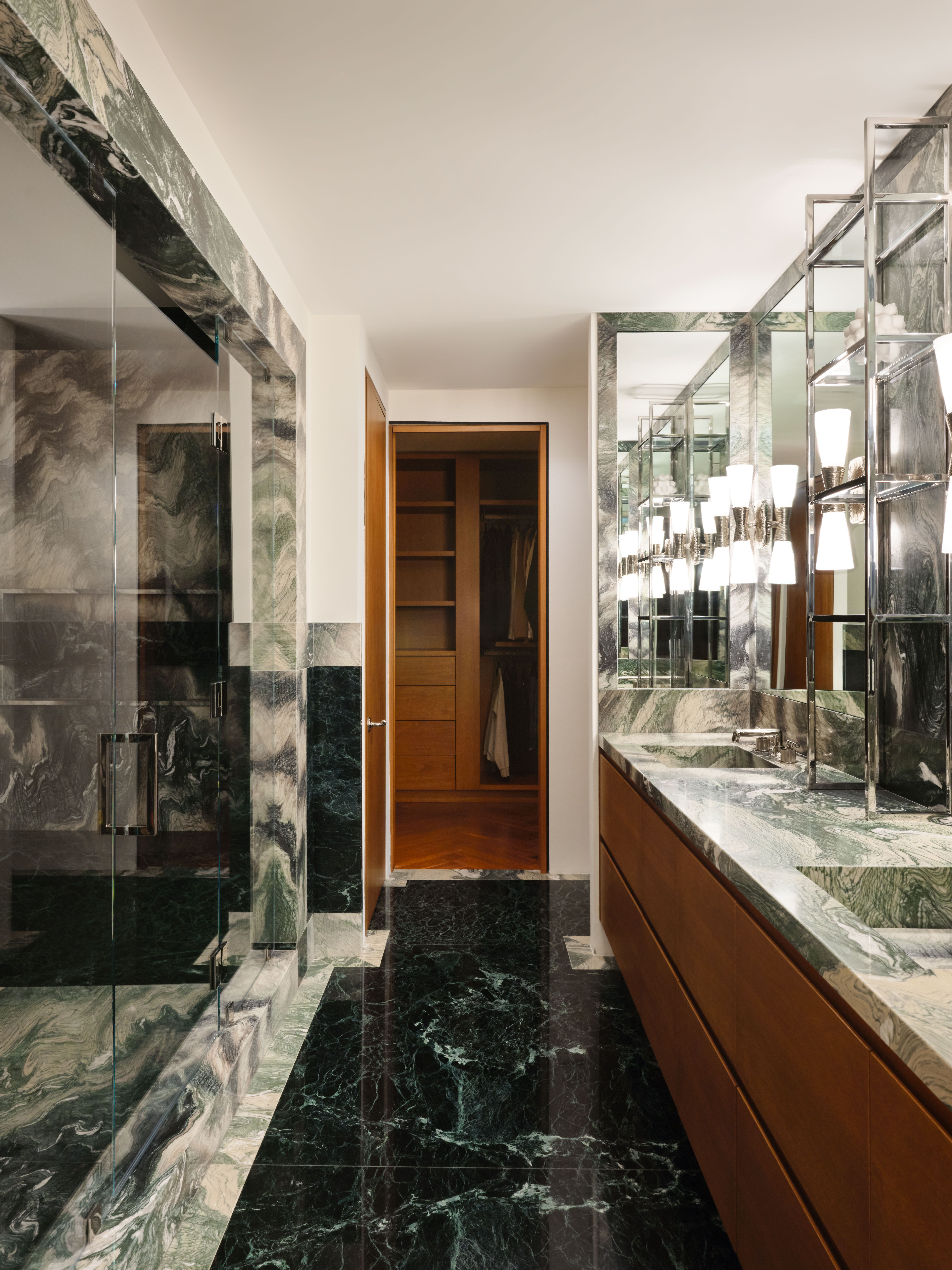 View of a black and green-tinted marble-clad bathroom decorated with bright lighting, wooden accents, and mirrors, leading into the closet
