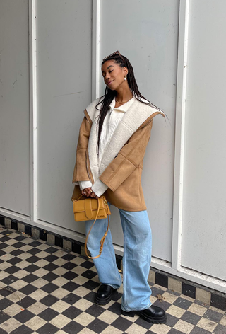 what to wear in 30-degree temperatures shown in a photo of a woman standing in a hallway wearing a shearing jacket over a white jumper with baggy jeans and black chelsea boots