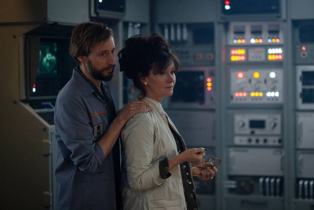 Still from the TV show Alien: Earth. A man is standing behind a woman and has his arm on her shoulder in a comfortable and familiar manner. They are standing in a room full of different computers.