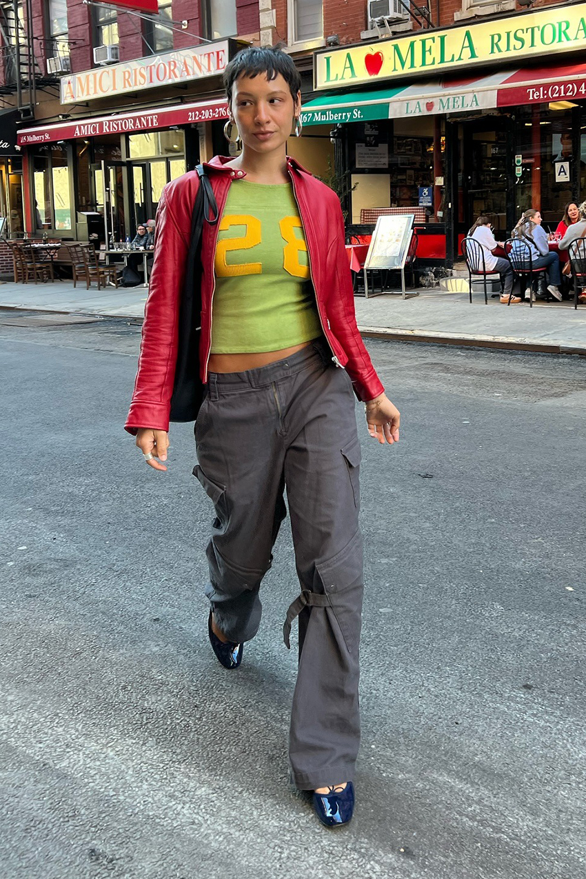 A woman wearing a red leather moto jacket, green and yellow jersey shirt, gray cargo pants and dark blue flats