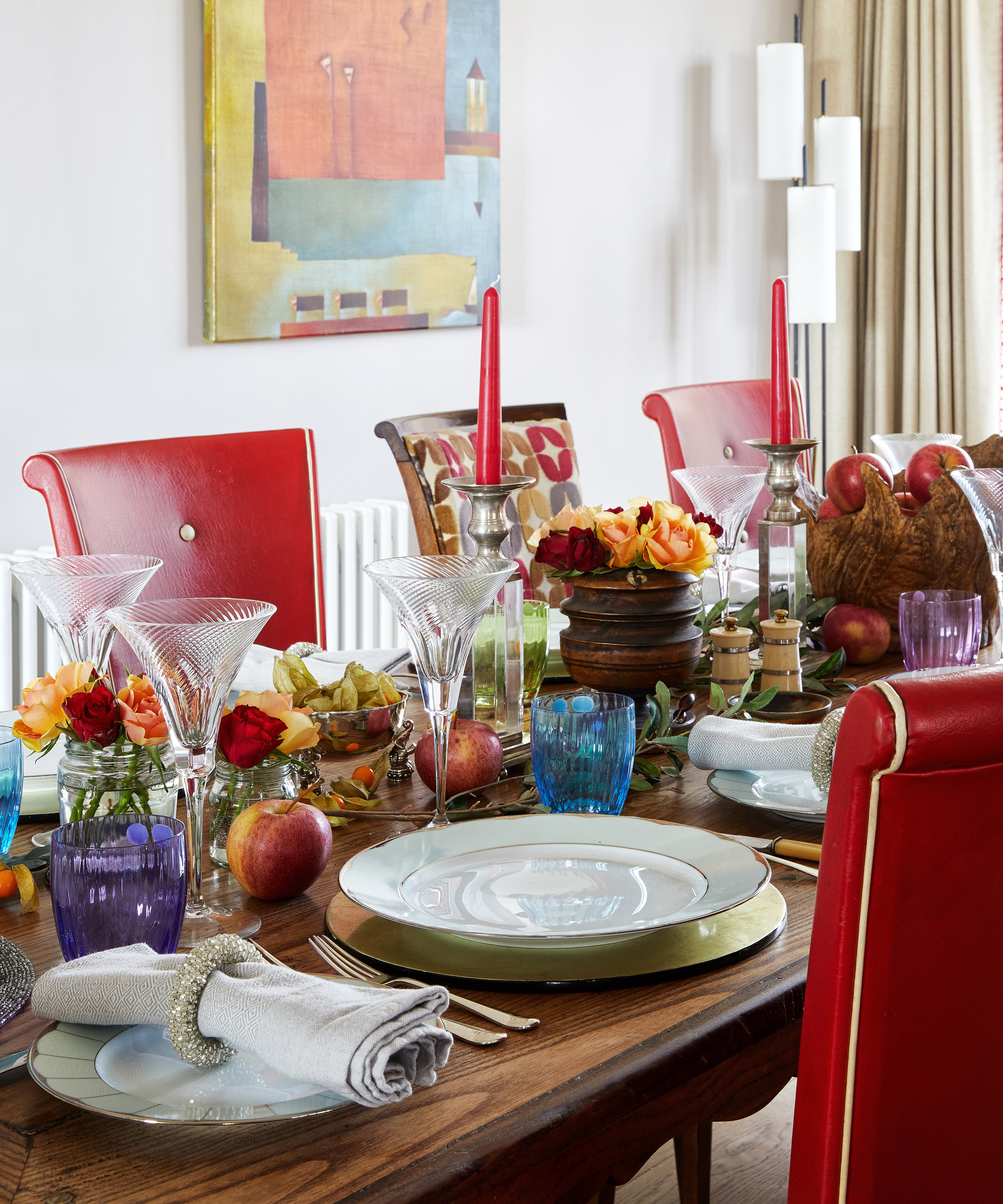 A dining table decor idea with red chairs, blue and purple glasses and fruit decor including red apples and orange physalis