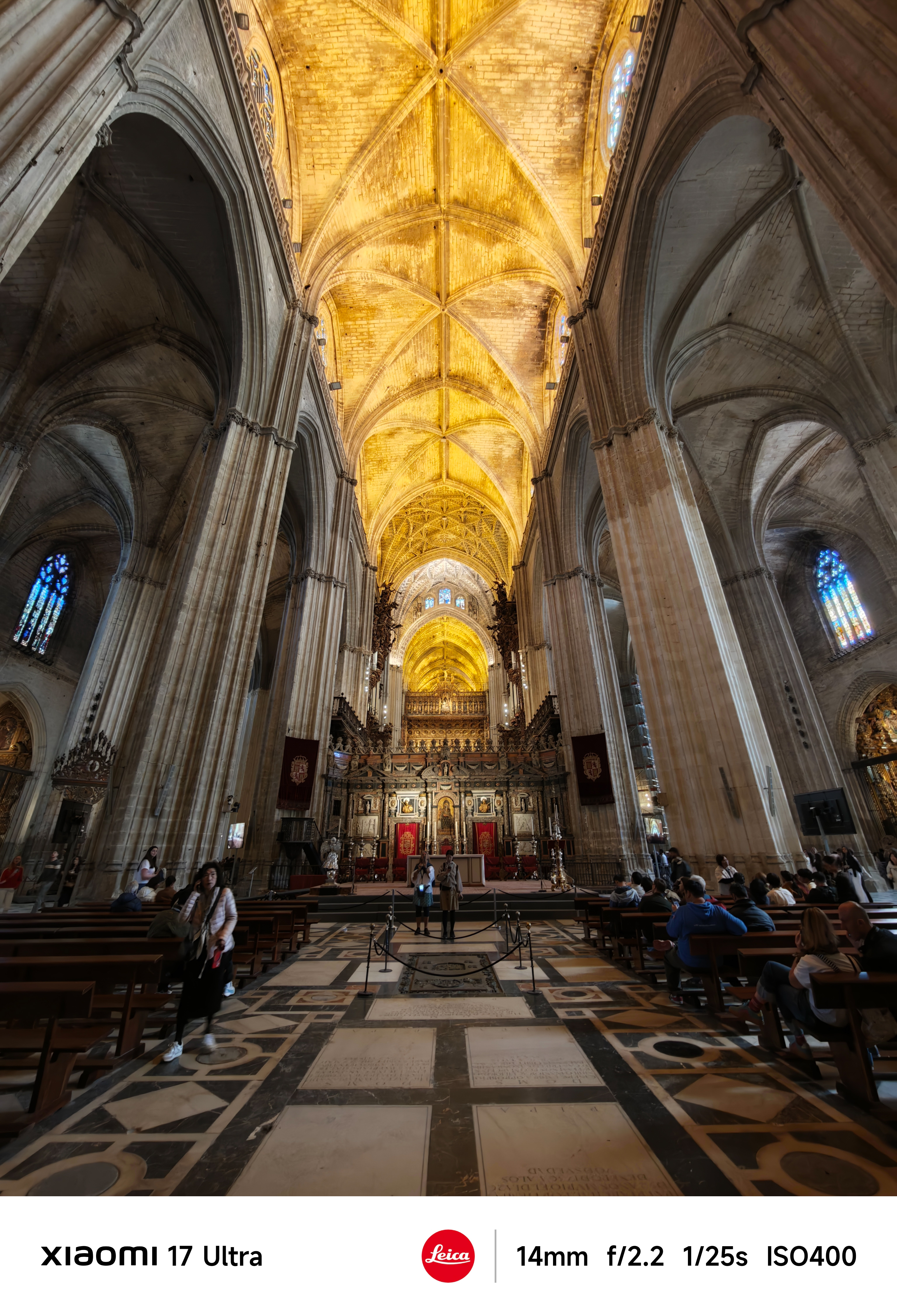 Interior of a grand cathedral nave with soaring vaulted ceilings glowing gold, stone columns lining both sides, and visitors seated in pews.