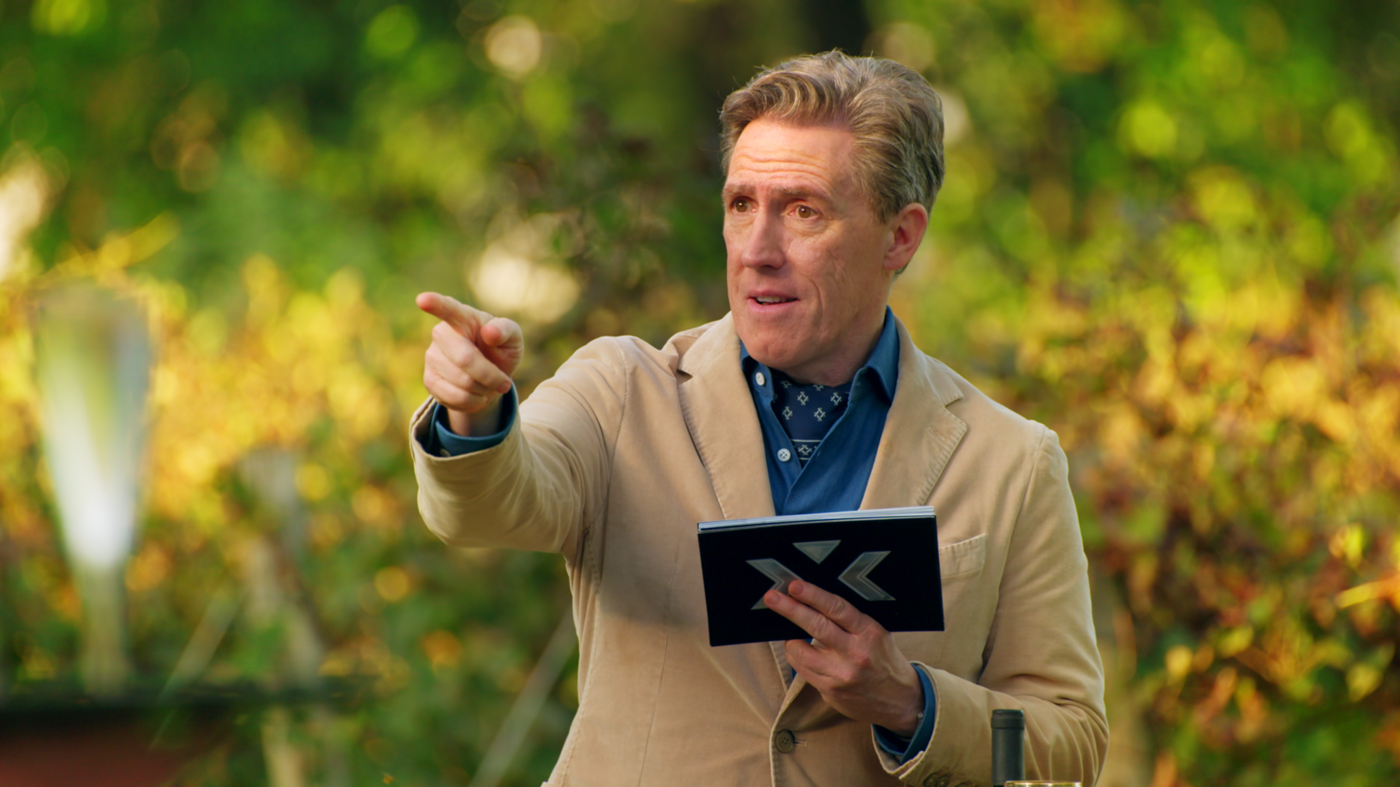 Rob Brydon standing in a garden with prompt cards in his hand, pointing at someone off camera.