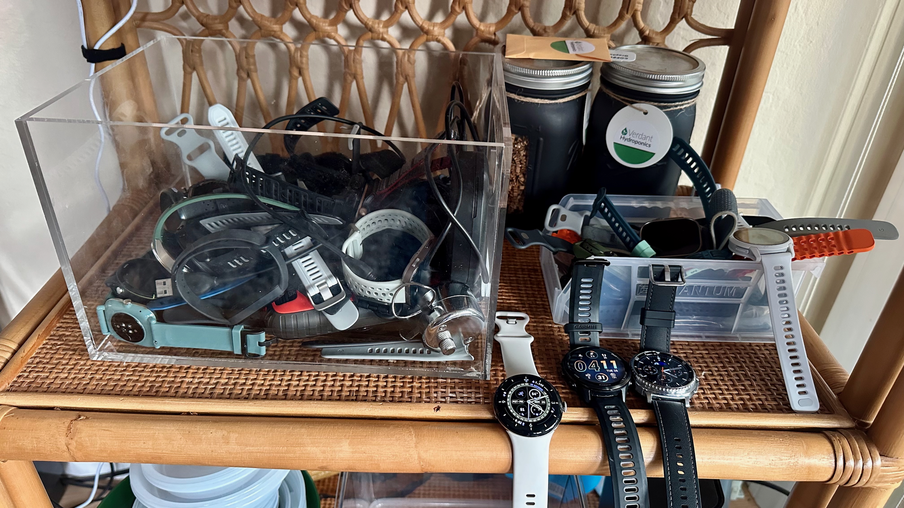 A photo of two plastic containers, one large and one small, sitting on a wooden bookshelf, both full of smartwatches, with three smartwatches (the Pixel Watch 4, Garmin Forerunner 970, and Galaxy Watch 8 Classic) sitting in front of both containers on the shelf.