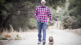 an older man smiles as he walks a small dog along a country road