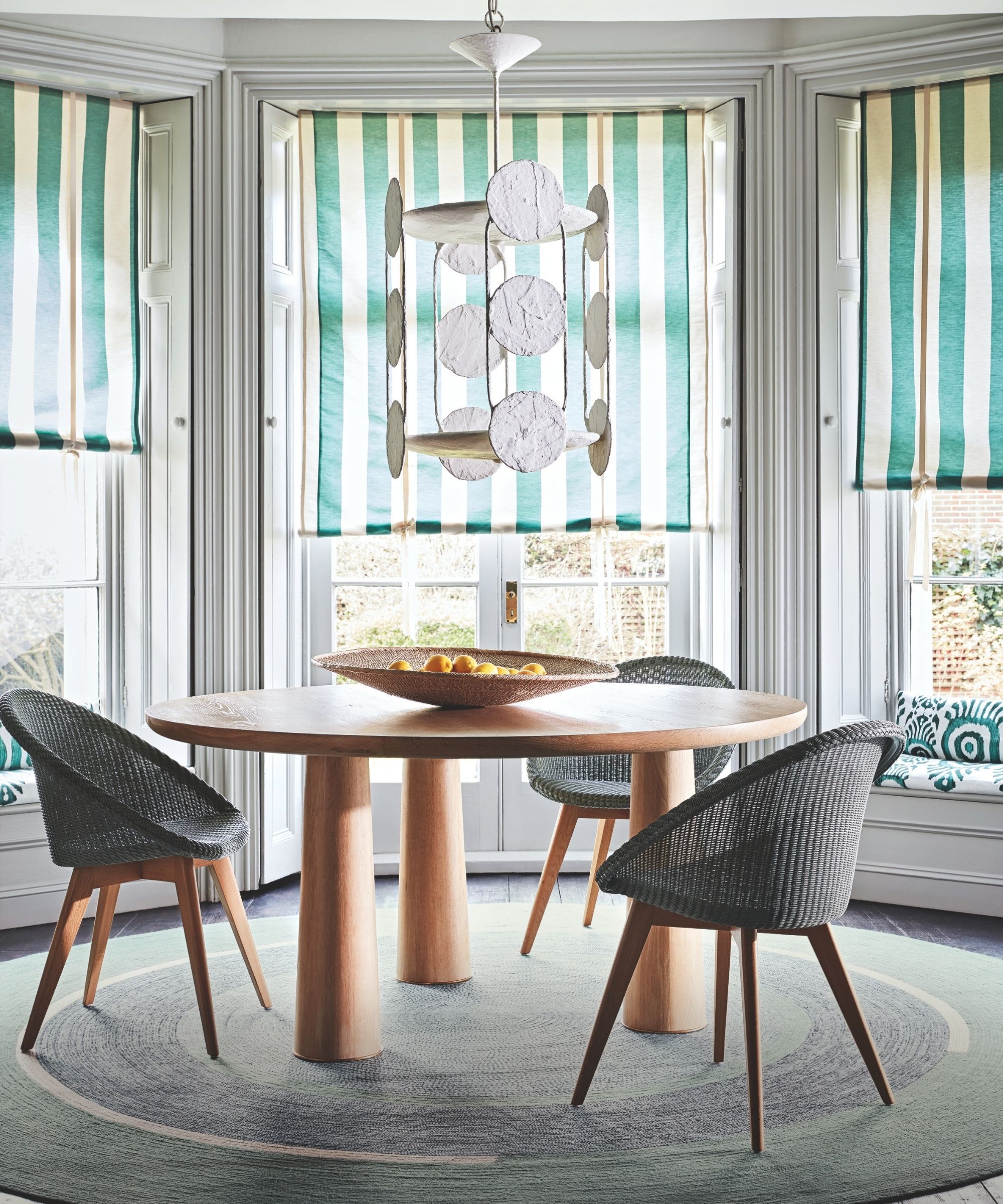 A large circular dining table with chairs, a circular rug, fruit bowl, striped blinds, and large bay windows