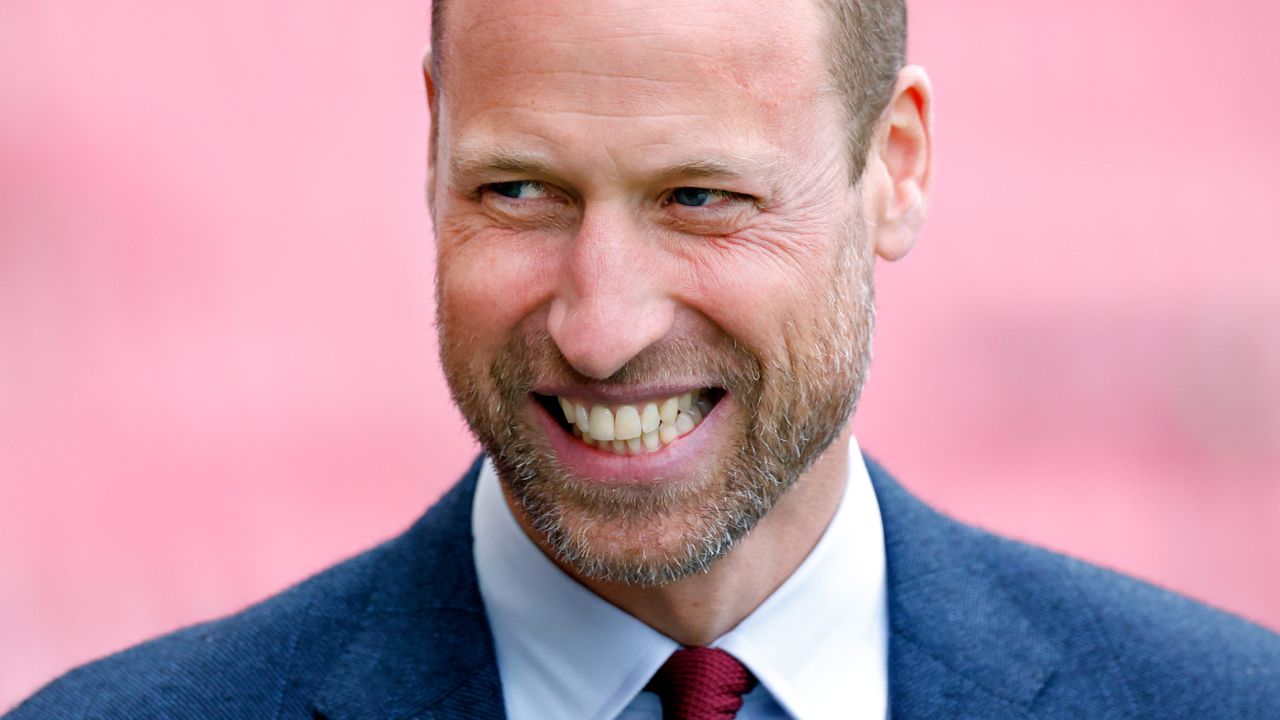 Prince William laughs, and has a rugged beard, while wearing a blue suit with a white shirt and red tie 