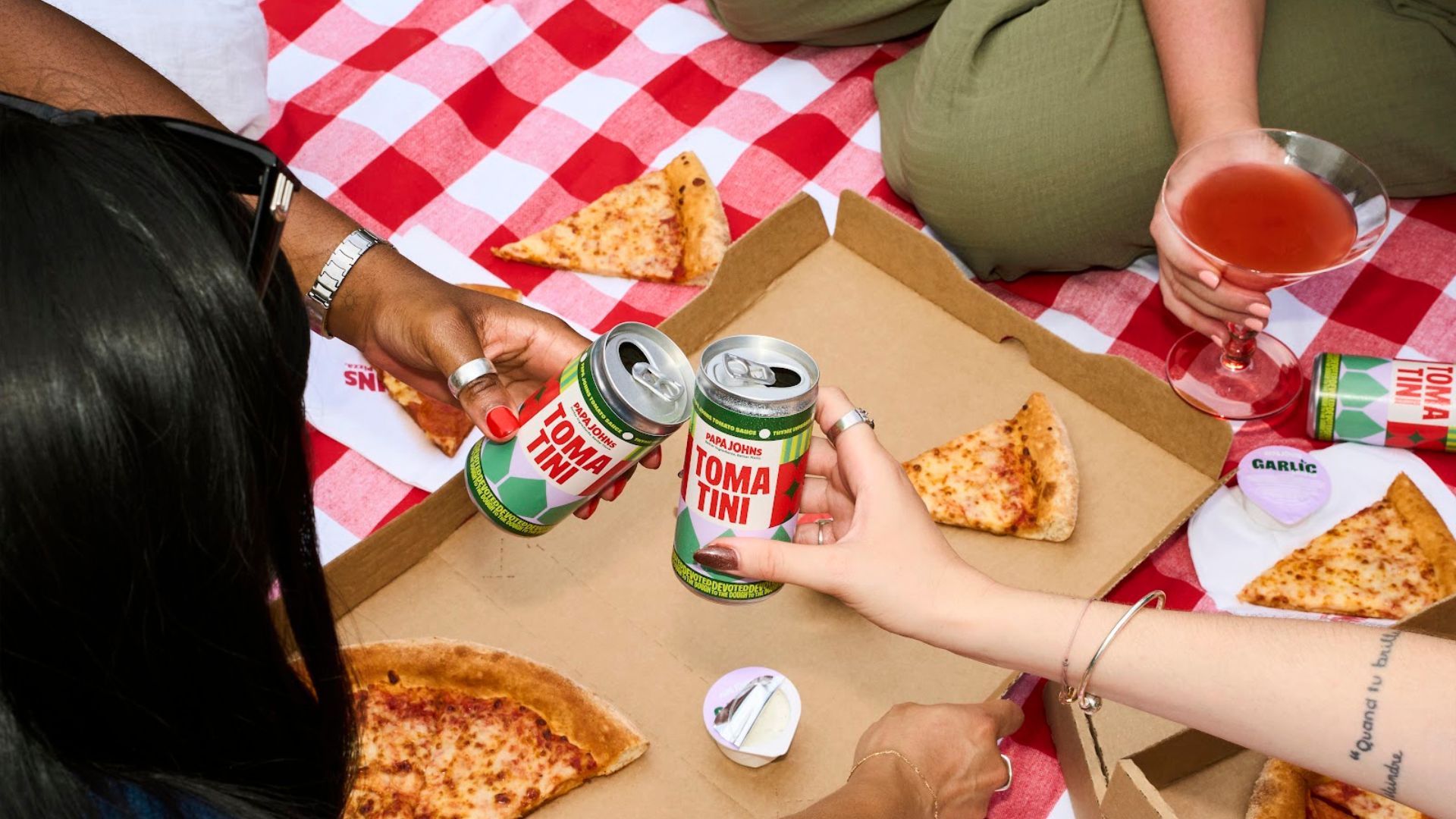 an image of the tomatini tinny. Two people are holding one each, and cheers-ing in the centre. They are sitting on a picnic blanket out of shot, and there is takeout pizza on the red gignham blanket. 