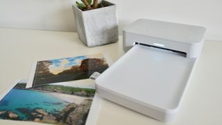 En vit HP Sprocket Studio står på ett vitt bord bredvid några utskrivna bilder och en liten krukväxt.