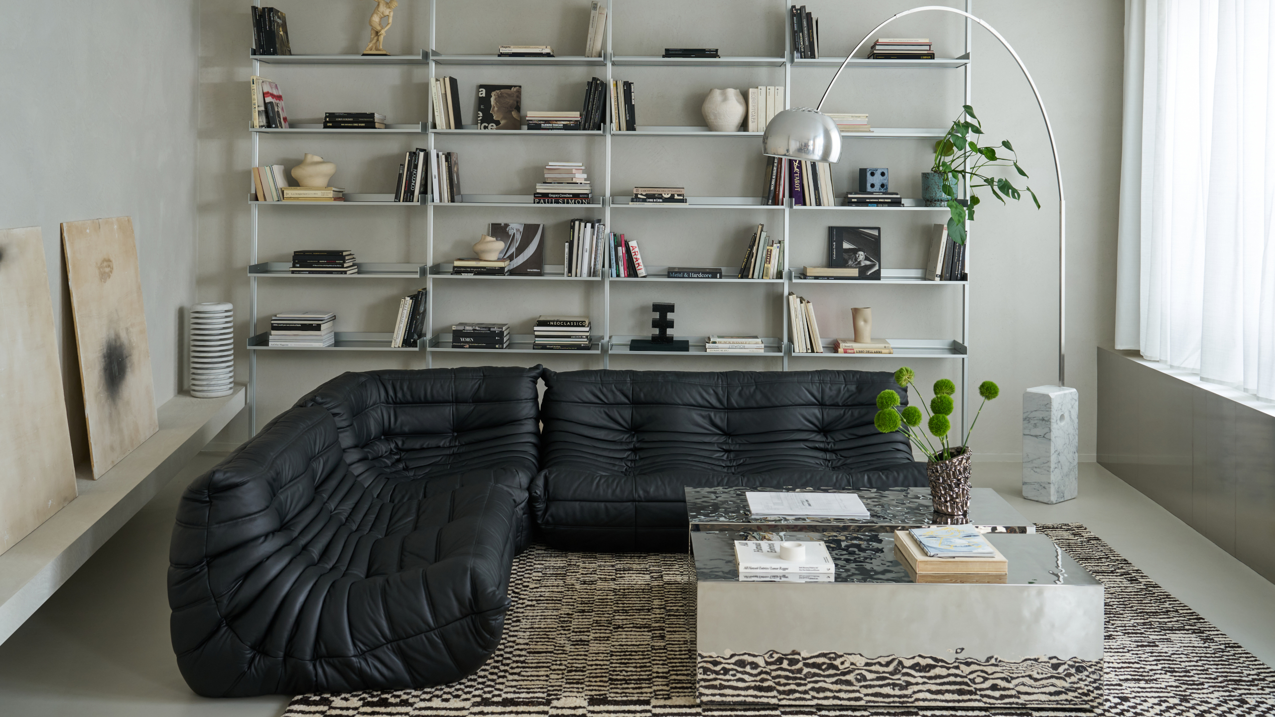 Living room with pale gray walls and steel panel, grey wall shelving, black Togo sofa, chrome square coffee table, black and white checkerboard rug and chrome curved floor lamp