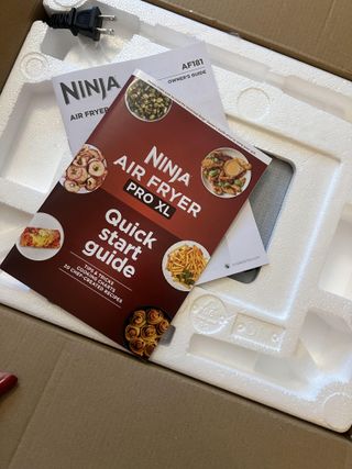 Shot from above, a picture of the open box of Ninja Air Fryer Pro XL, revealing plastic molded protective styrofoam and quick start guide which is a red booklet