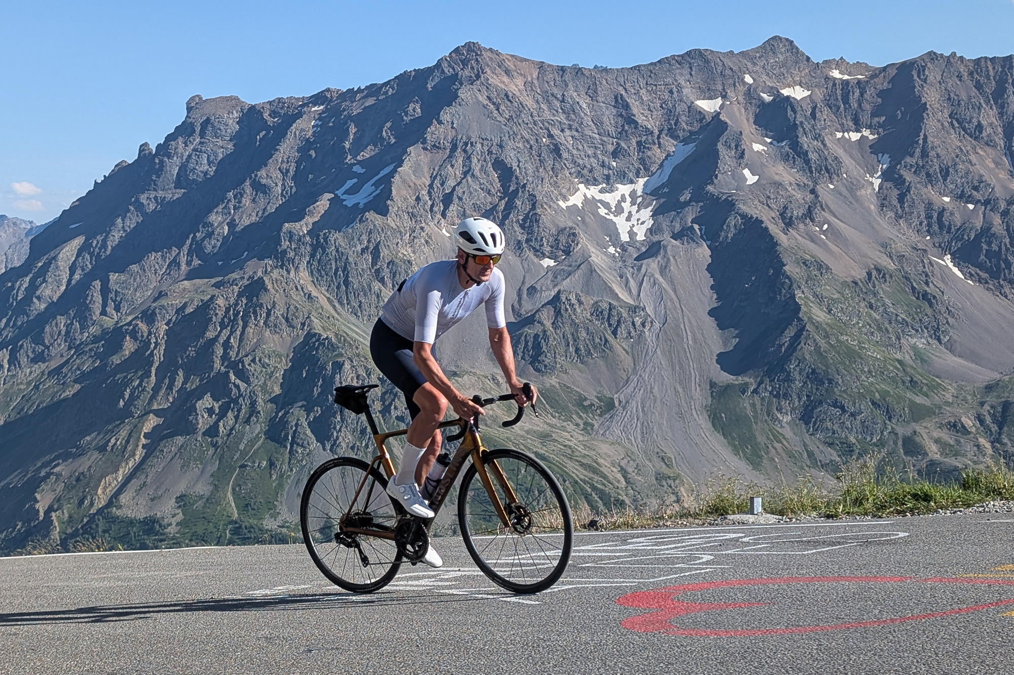 Man riding an orange bike up a hill wearing blue shorts, grey top, white helmet and white shorts with a mountain background
