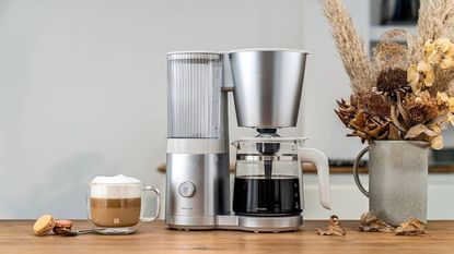 A stainless steel Zwilling drip coffee maker on a wooden countertop with a cappuccino, and a vase of dried flowers