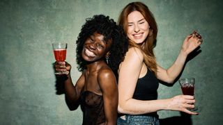 Two cheerful young multiracial friends standing back to back, smiling and holding drinks while enjoying a fun time during a lively party or social event