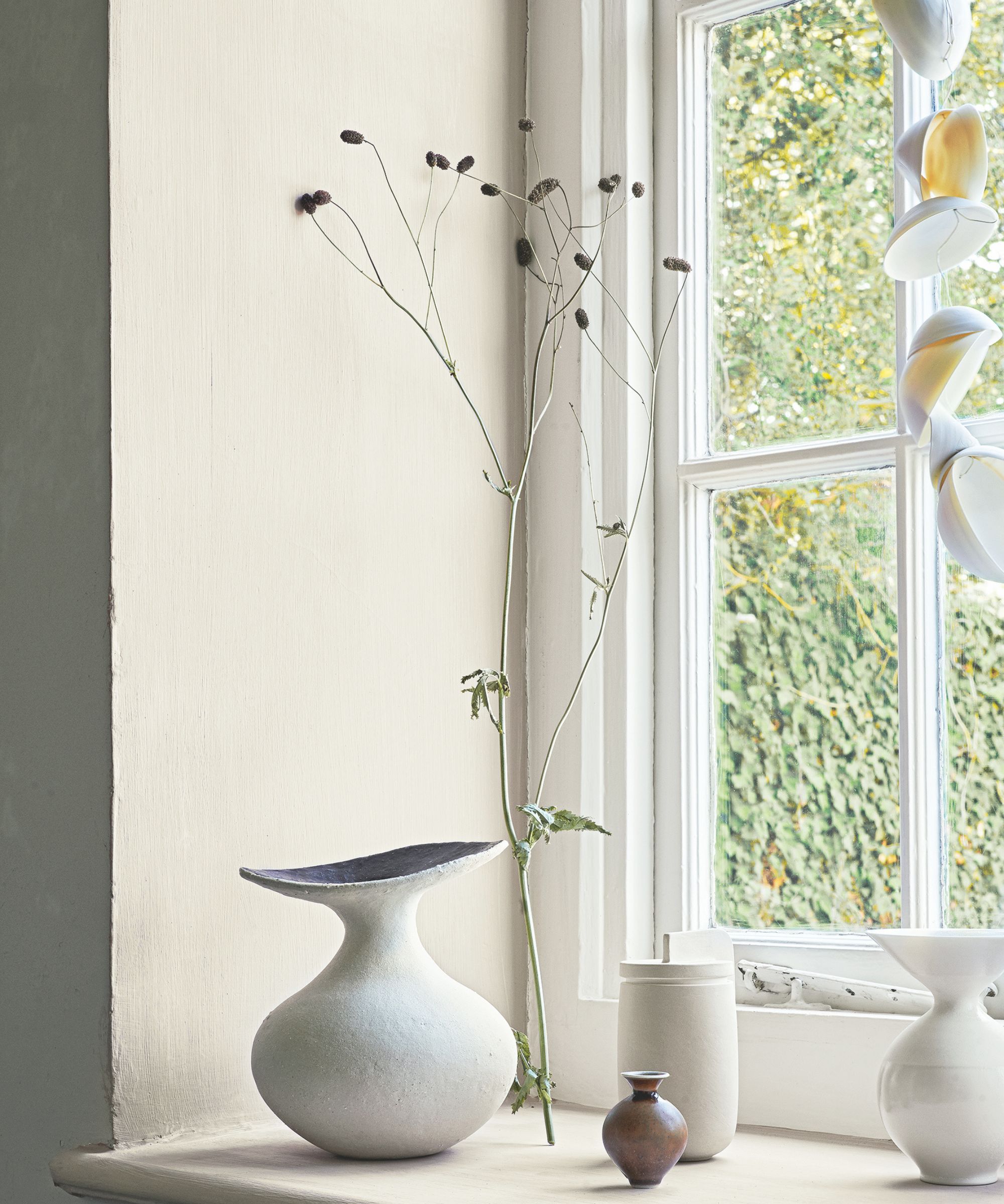A bright window with white walls, eclectic vases, dry flowers and white shells hanging on a string.
