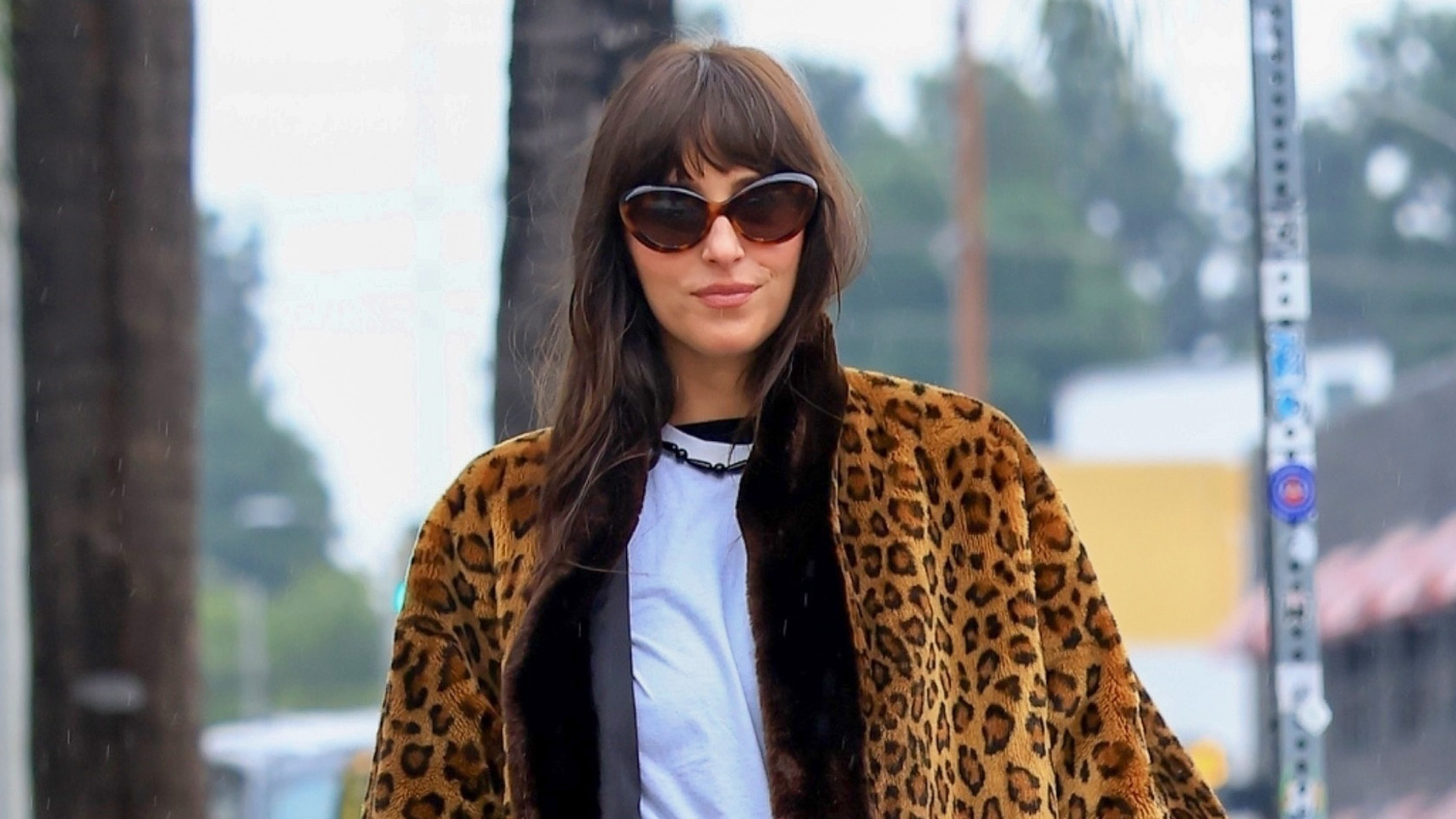 Dakota Johnson wearing a leopard fur coat, white T-shirt, jeans, and patent leather Adidas Samba sneakers