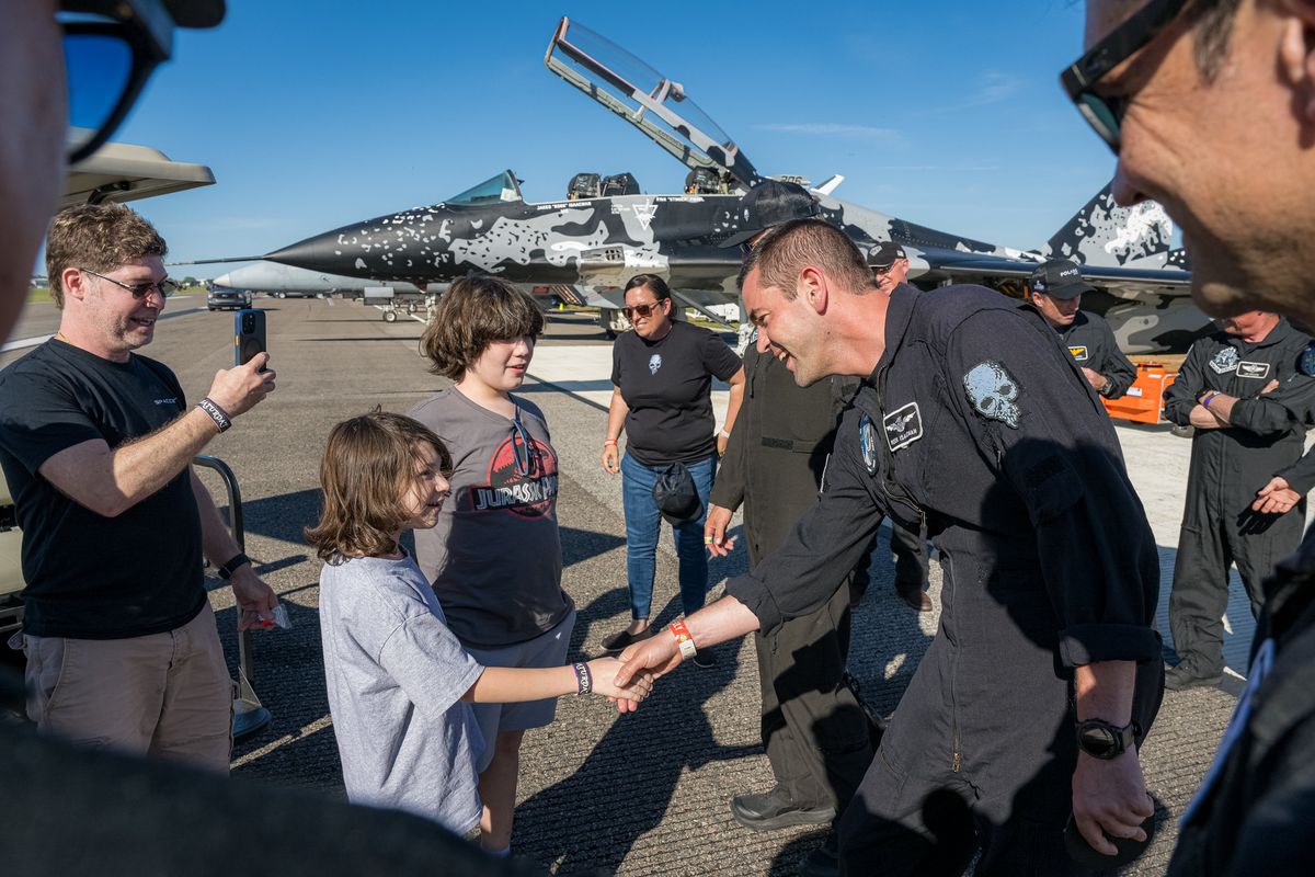 a man in a blue flight suit shakes hands with a child on a tarmac, with a fighter jet in the background