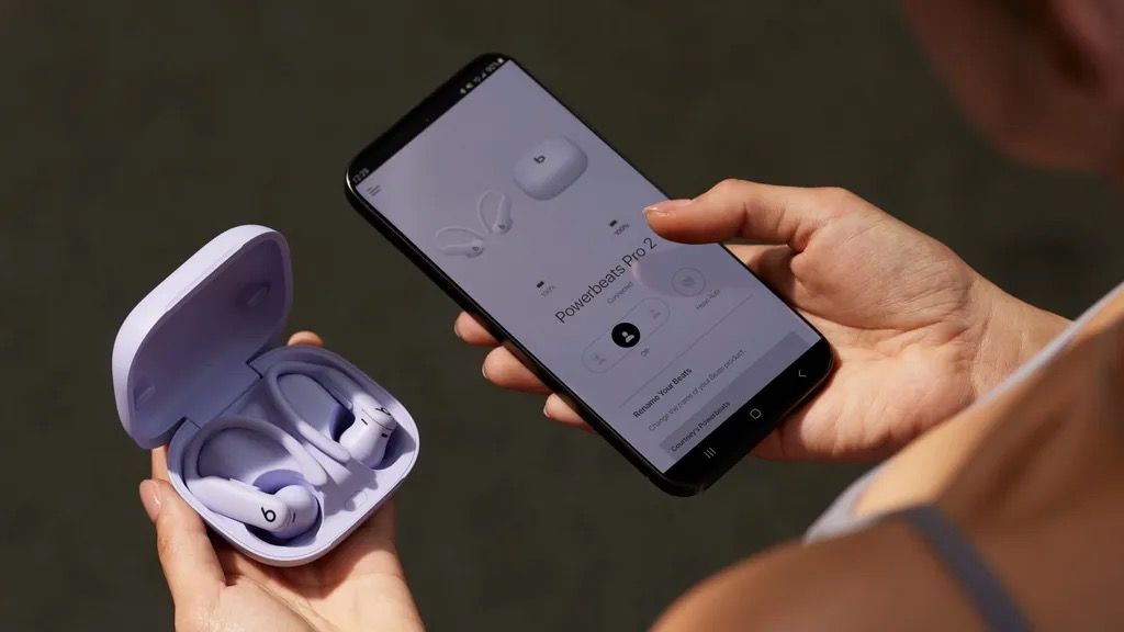 A pair of Powerbeats Pro 2 wireless earbuds in their case, held next to an iPhone with the Beats app on screen.