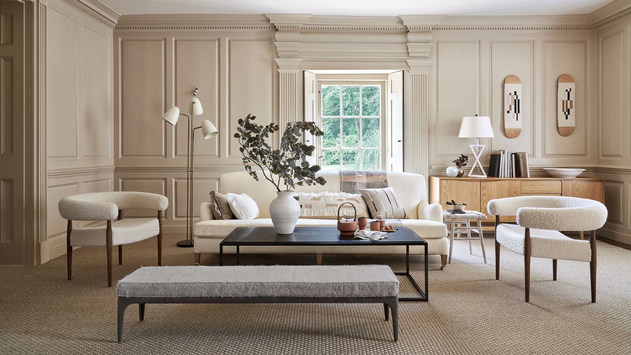 Beige painted living room with paneling around the room, spacious seating area with cream sofa and two armchairs in boucle fabric, black rectangular coffee table and grey footstool, white metallic floor lamp, window with shutter, lights wood sideboard with white table lamp and accessories 