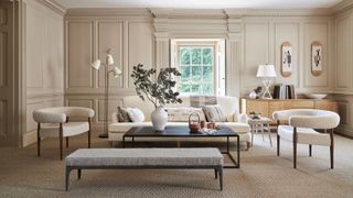 Beige painted living room with paneling around the room, spacious seating area with cream sofa and two armchairs in boucle fabric, black rectangular coffee table and grey footstool, white metallic floor lamp, window with shutter, lights wood sideboard with white table lamp and accessories