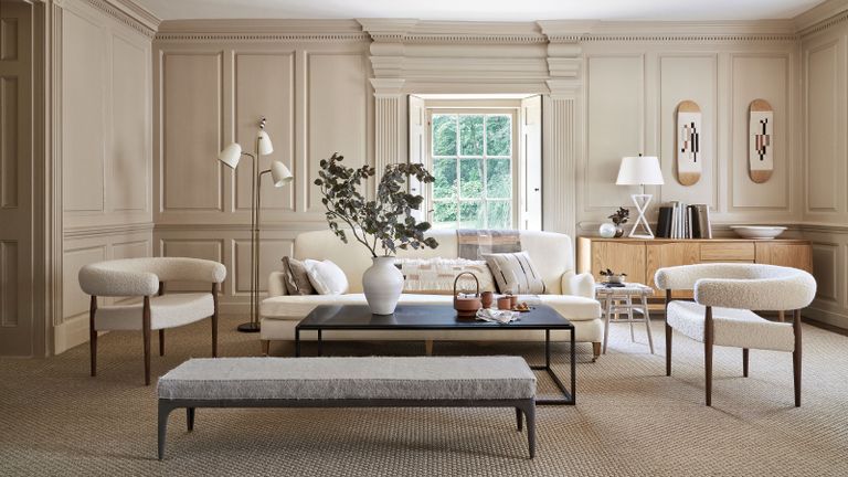 Beige painted living room with paneling around the room, spacious seating area with cream sofa and two armchairs in boucle fabric, black rectangular coffee table and grey footstool, white metallic floor lamp, window with shutter, lights wood sideboard with white table lamp and accessories 