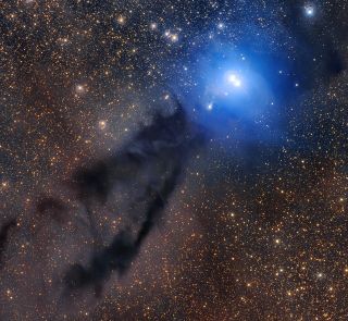 a huge glow of blue stars is visible in a telescopic image. a streak of dark dust runs from the blue stars to the bottom left of the image