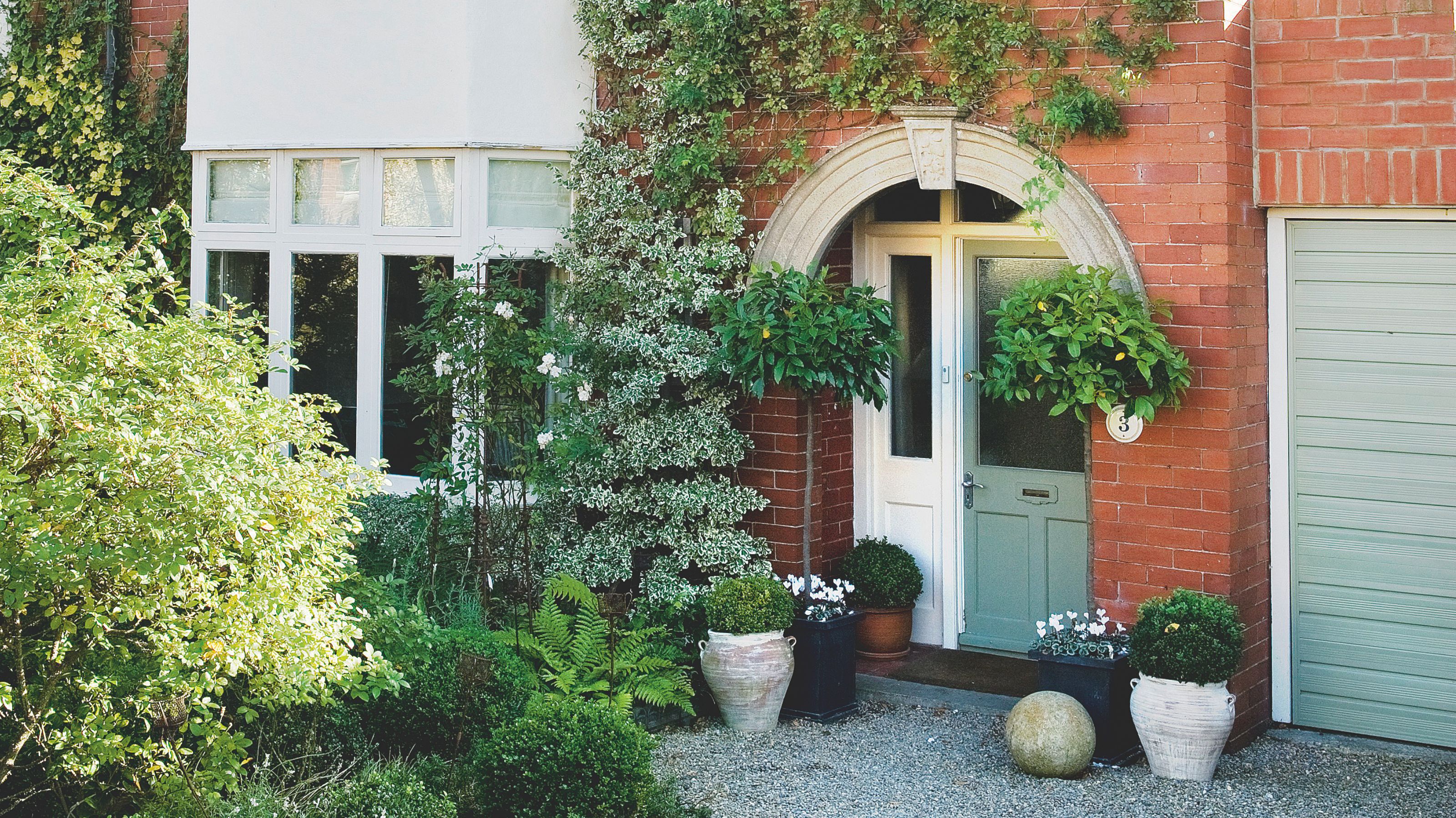 Exterior of brick house with matching colour front door and garage door surrounded by established plants and pretty pots