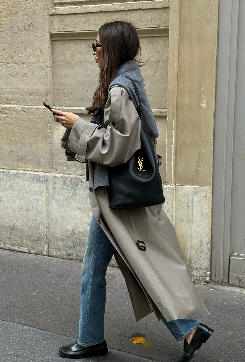 A woman walking down the street wearing a trench coat with a gray sweater around the shoulders, straight jeans, black loafers, and a black slouchy bag.