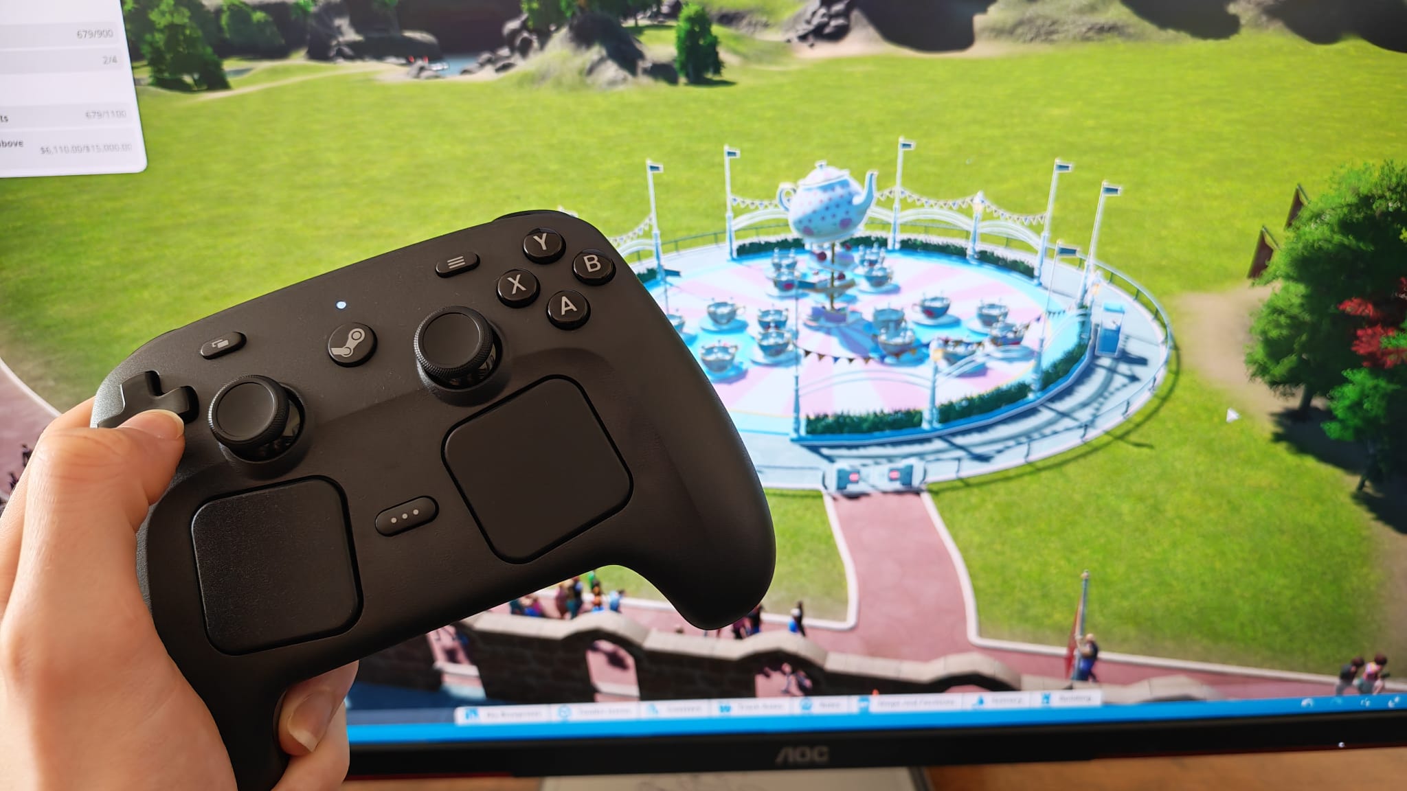 A Steam Controller being held up in front of a gaming monitor playing Planet Coaster