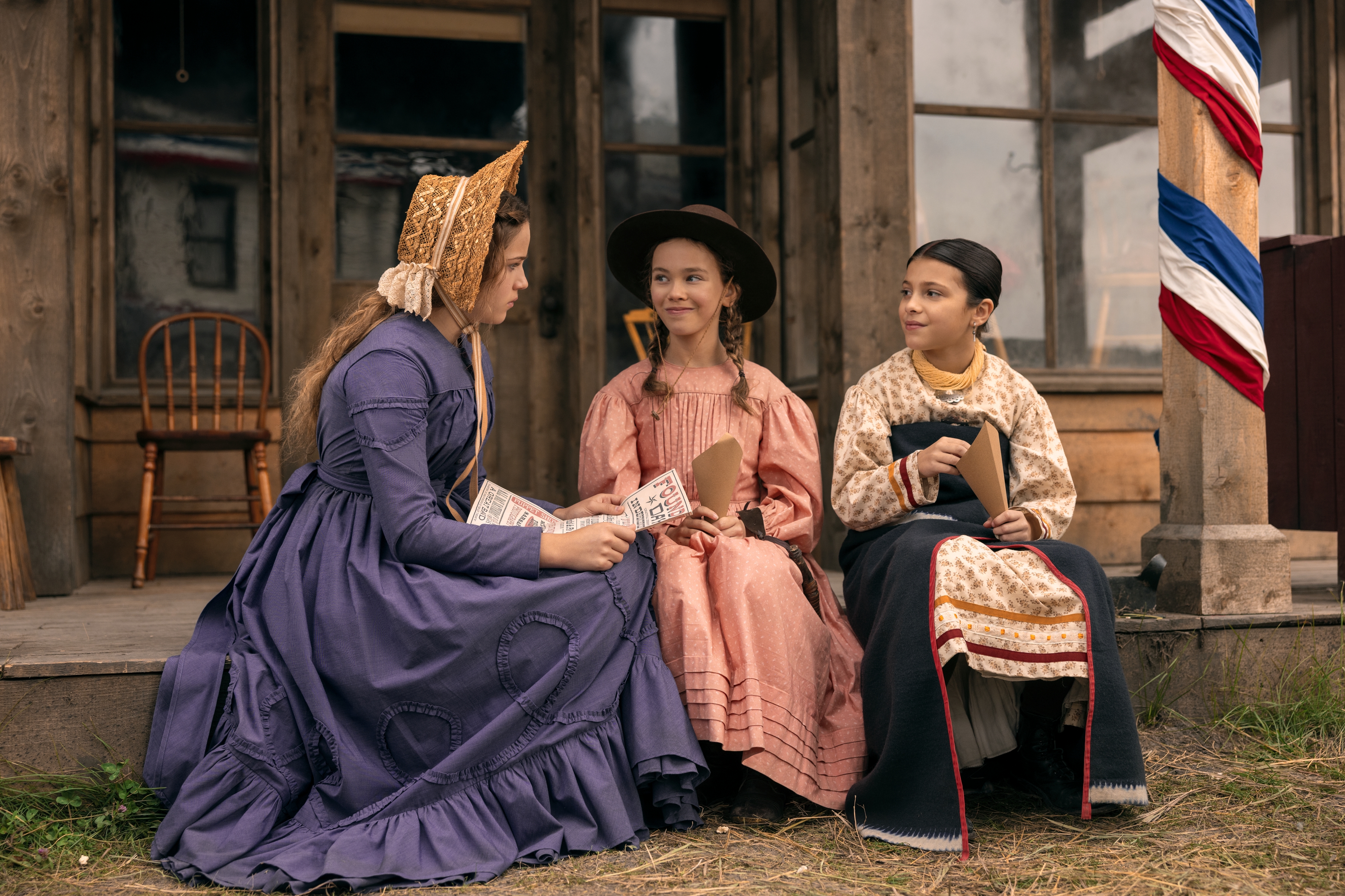 Skywalker Hughes as Mary Ingalls, Alice Halsey as Laura Ingalls, Wren Zhawenim Gotts as Good Eagle sit on the front step of a frontier-era building in Netflix's 'Little House on the Prairie.'