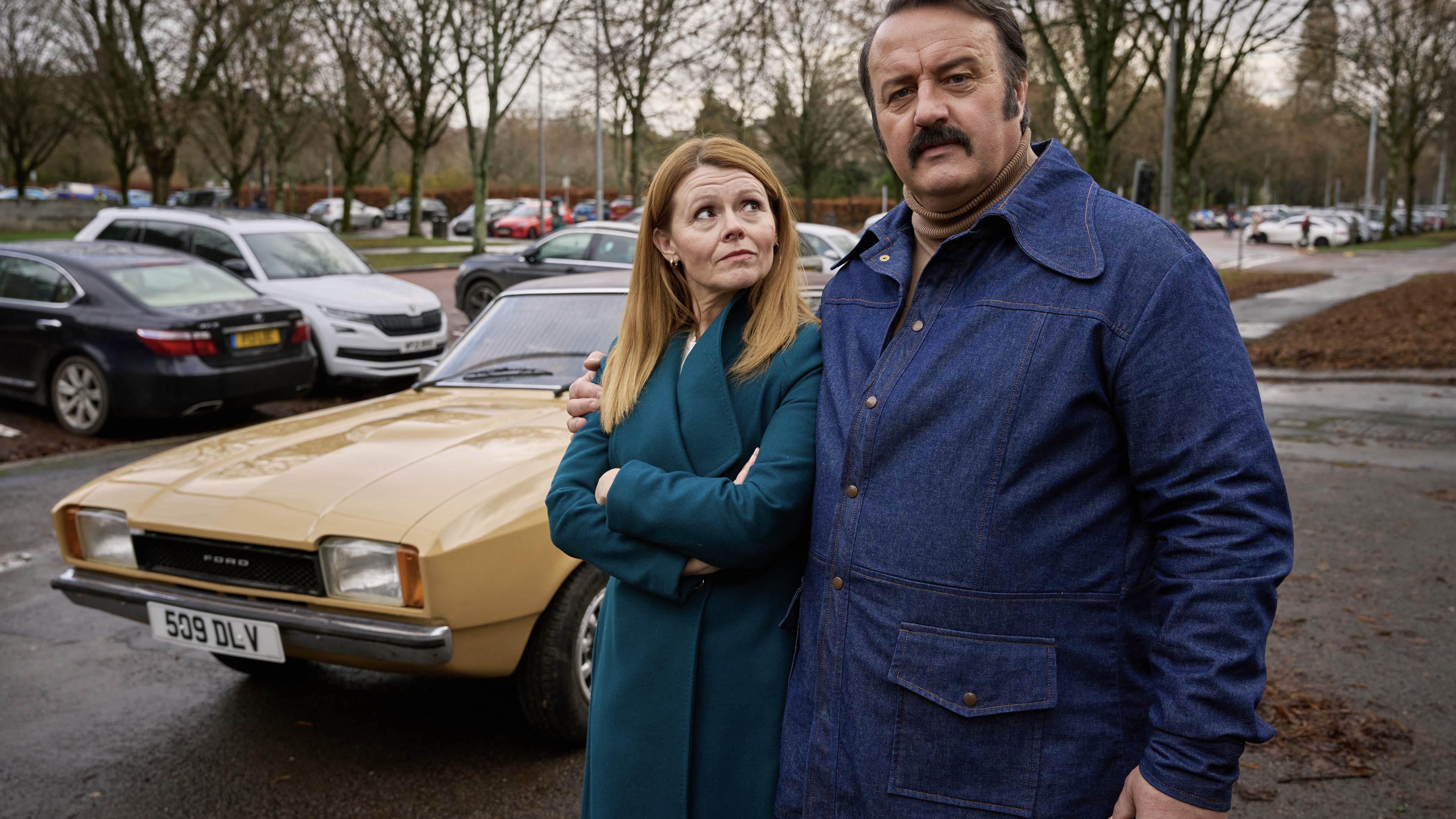 Sian Gibson in a green coat as Mel stands next to Mike Bubbins in a denim jacket as Tony Mammoth alongside a beige Ford in Mammoth.