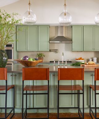 Mint green kitchen cabinets with matching base and wall cabinets, tan and black bar stools, fluted glass pendant lights, vase with foliage, hardwood floor