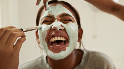 A woman in laughing while her friends (hands shown) are applying a green clay mask to her face