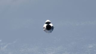 a white space capsule approaches the international space station, with a few clouds over the ocean in the background