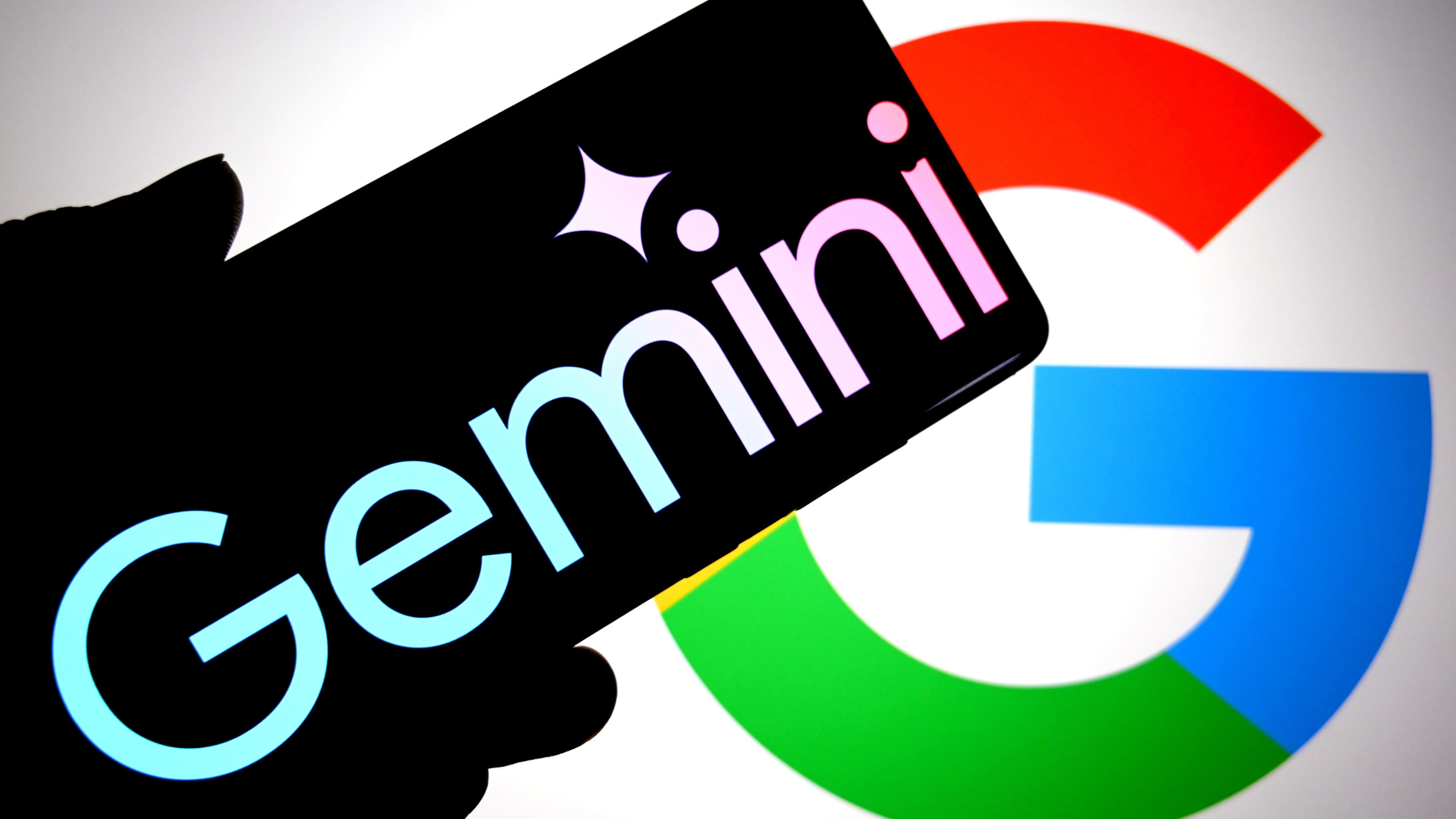 A Google Gemini logo is seen displayed on a smartphone with a Google logo in the background.