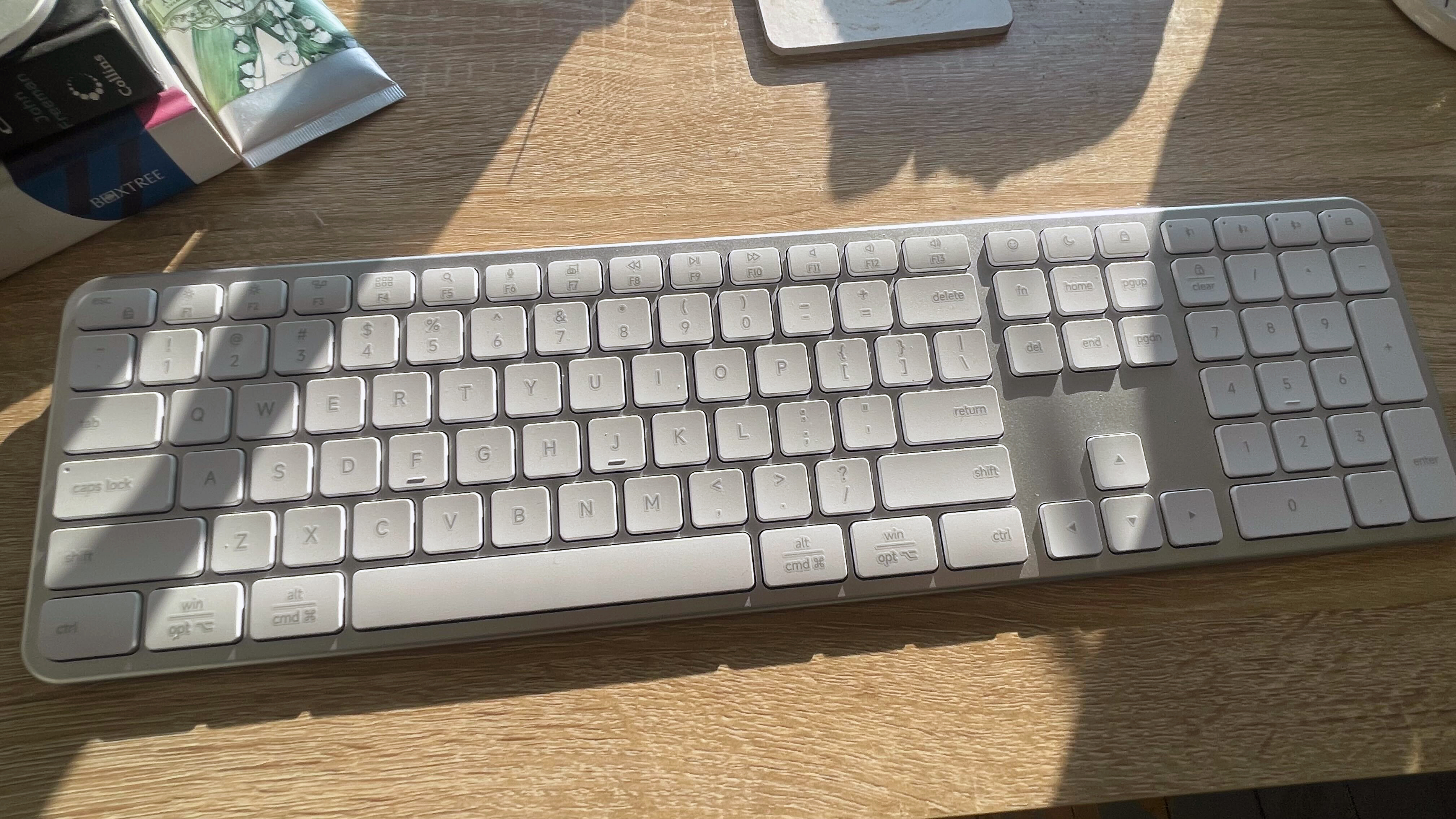A silver grey Satechi Slim EX3 keyboard and Slim EX mouse on a wooden desk with sunlight shining on it from a window in the background