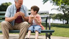 A grandpa and grandson share a sandwich on a park bench.