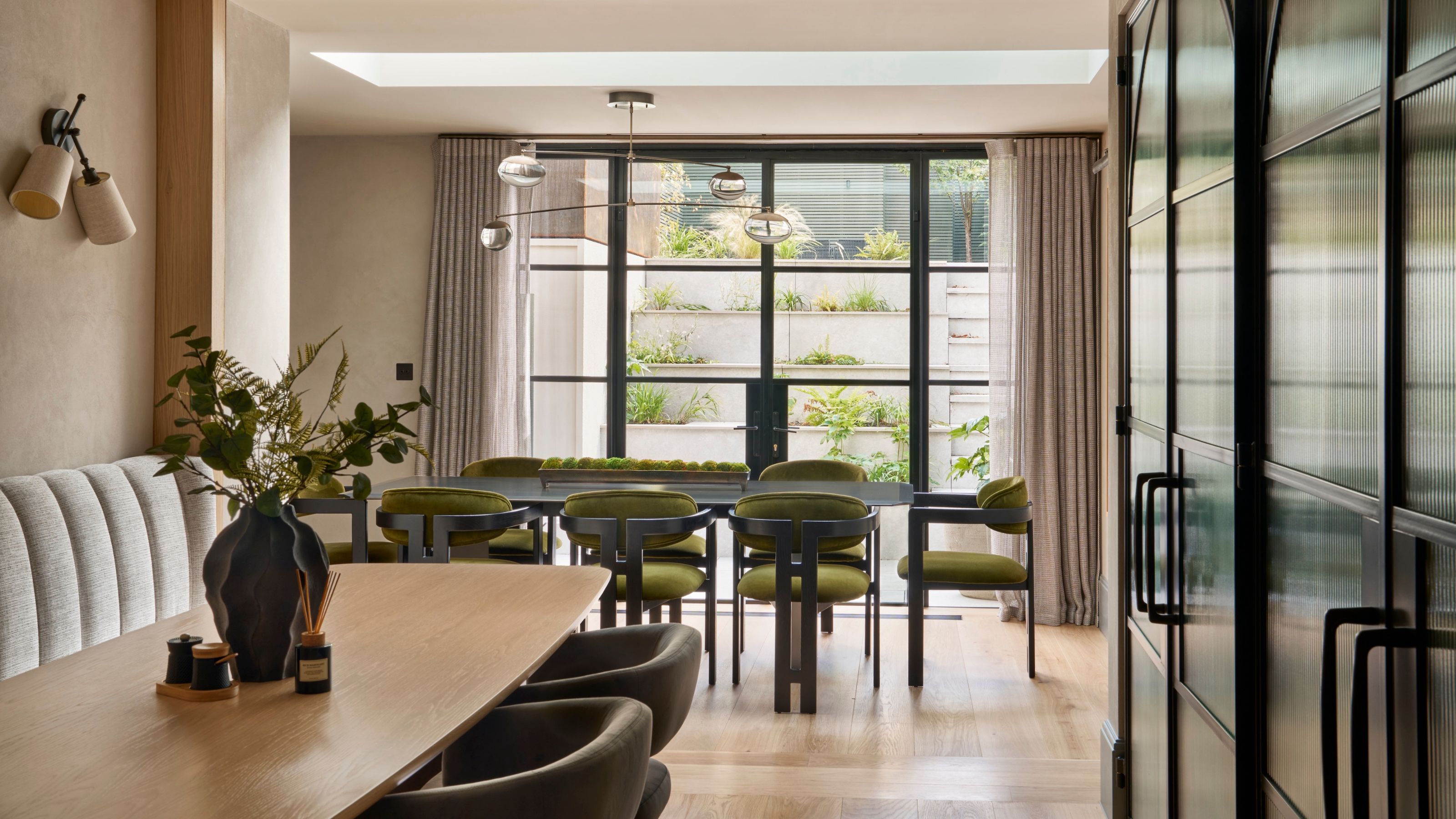 A banquette seating area in front of dark built-in cabinetry with a dining table facing the opposite way at the other end of the room in front of glass doors leading to the garden