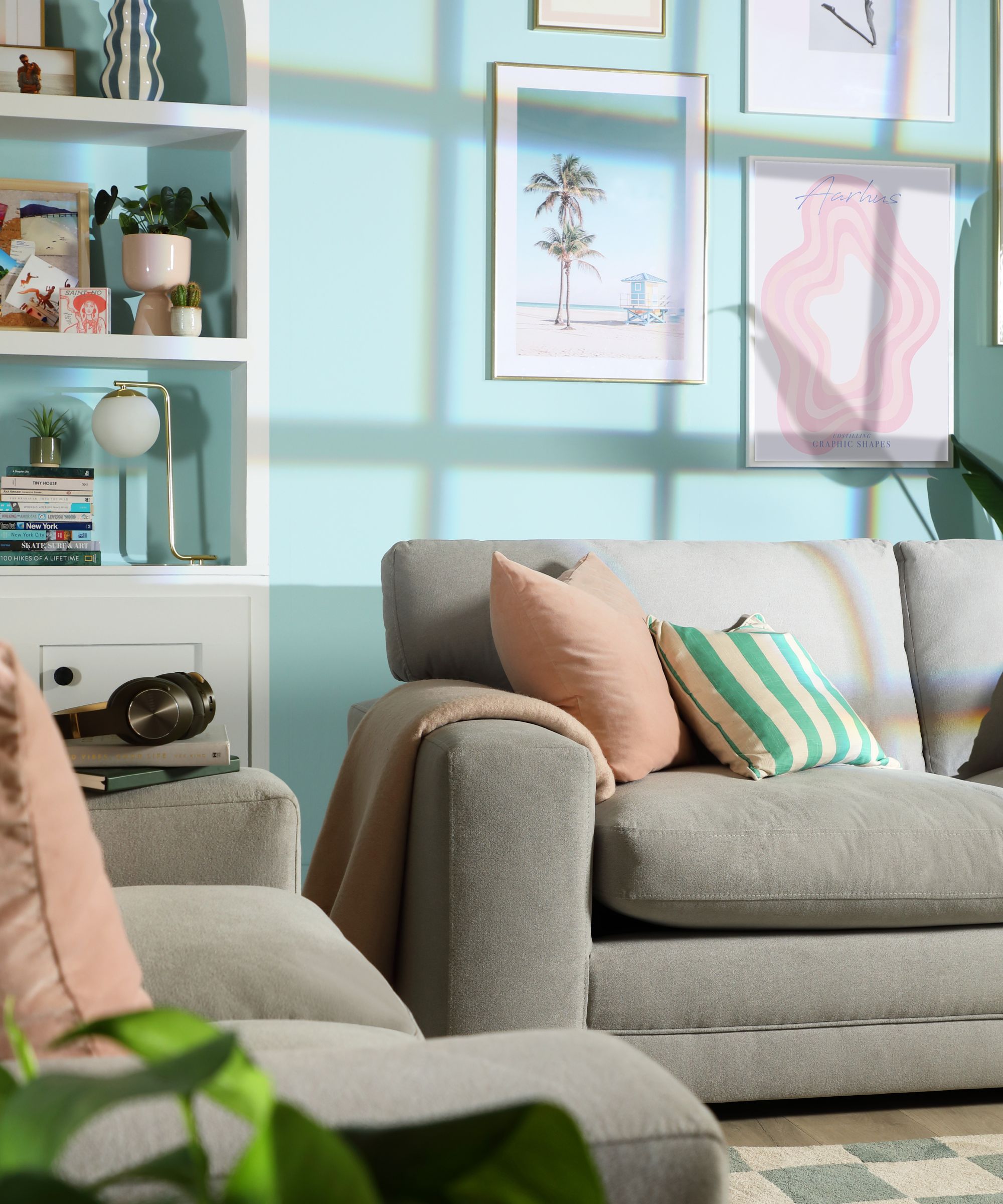 A grey upholstered sofa with pink and striped cushions in a mint green living room with a gallery wall of framed prints, built-in shelving and a checked rug