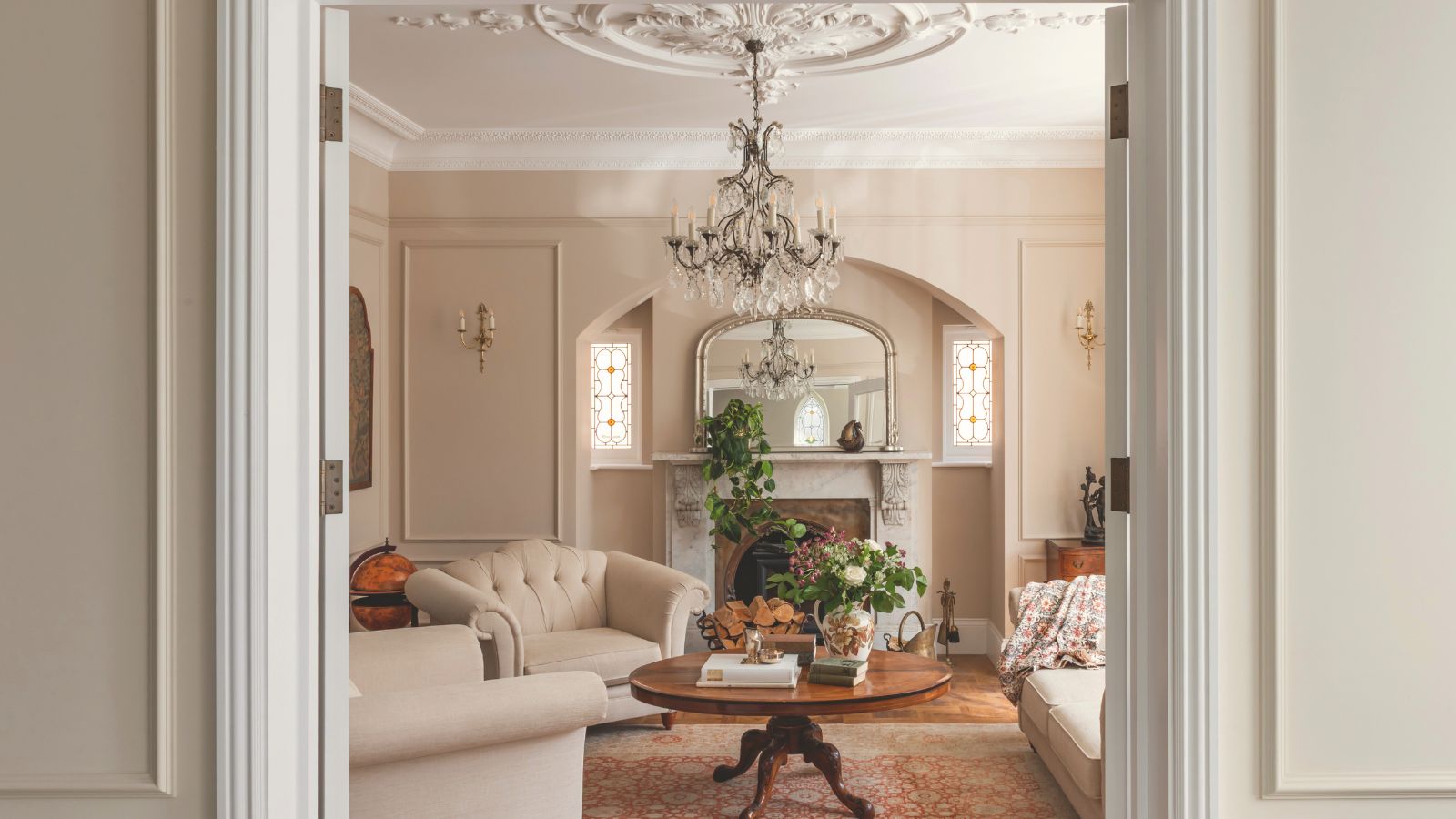 A traditional cream living room, with cream upholstered arm chairs around a round wooden coffee table. A crystal chandelier hangs rom a ceiling rose 