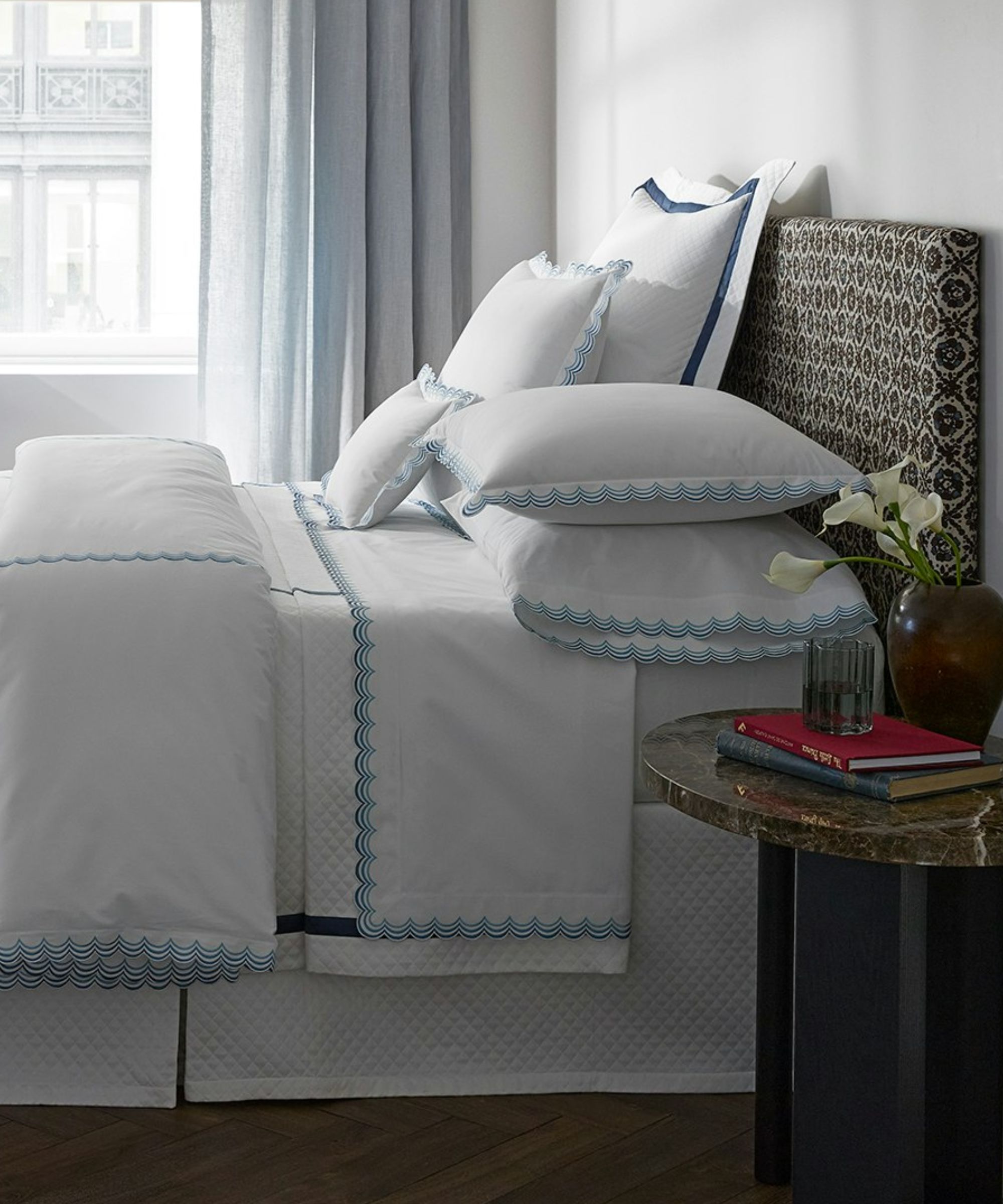 Modern bedroom with blue and white bedding, black and white patterned headboard, marble bedside table with vase and books, blue curtains