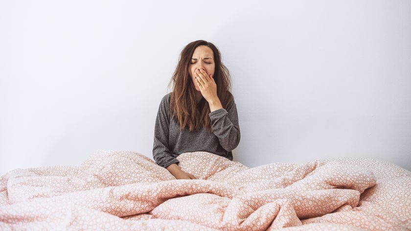 A woman with long brown hair sits up in bed covered in a pink comforter and yawns because she is so sleep deprived