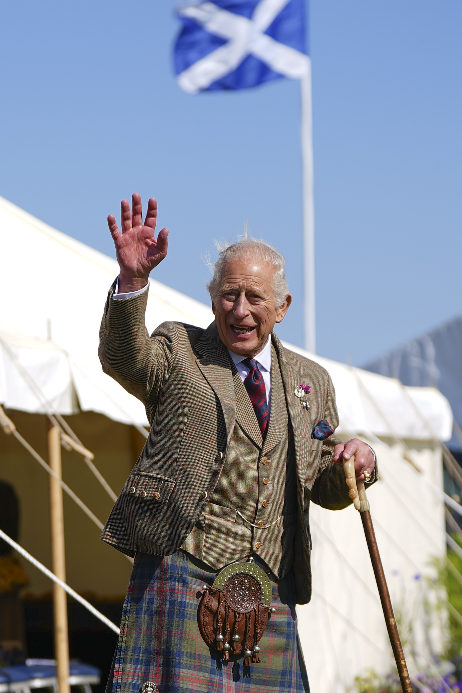 King Charles dressed in a kilt and brown blazer, waving in front of a Scottish flag